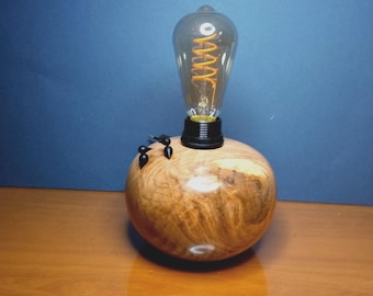 May include: A table lamp featuring a spherical, polished light-brown wooden base with visible natural grain patterns. A black socket is mounted on top, holding a clear glass Edison-style light bulb with a visible glowing orange spiral filament.