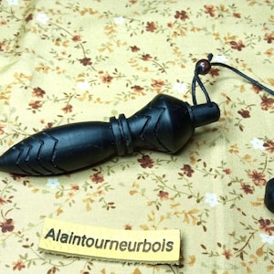 May include: A black wooden pendulum with a pointed end and carved details, suspended by a black cord. The pendulum is lying on a floral patterned fabric. The text "Alaintournebois" is on a small label.