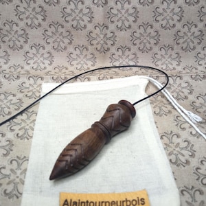 May include: A dark brown wooden pendulum with carved chevron patterns, on a white linen pouch. A black cord is attached to the pendulum. The wooden tag below reads "Alaintourneurbois".
