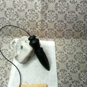 May include: A black wooden pendulum with a smooth, tapered shape attached to a thin black cord. It rests on a small white fabric pouch featuring a tan leather label with the text Alaintourneurbois printed in black ink, next to a porous stone.