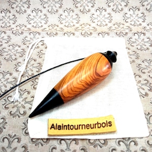 May include: A wooden plumb bob featuring a polished light brown wood grain body with dark brown pointed tips at both ends. A thin black cord is attached to the top. A small rectangular tan label with the text Alaintourneurbois is placed below.
