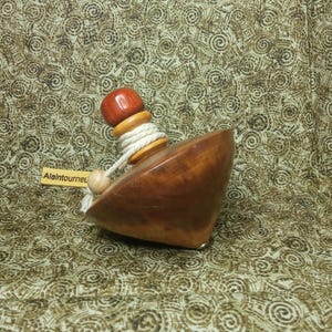 May include: A wooden spinning top with a rounded base and a red top. The top is attached to a white string and has a small wooden bead at the end. The text "Alaintourneu" is on a small tag.