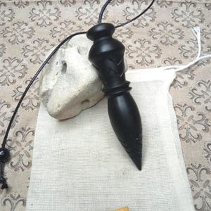 May include: A black wooden pendulum with a carved geometric pattern, suspended from a thin black cord. The pendulum rests next to a light-colored, porous stone and a small white fabric pouch featuring a tan leather tag labeled Alaintourneurbois.