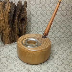 May include: A wooden inkwell with a glass ink pot and a wooden dip pen. The inkwell is made of light brown wood with a circular shape. The dip pen is long and thin with a brown wooden shaft.