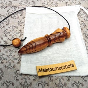 May include: A handcrafted wooden pendulum with a pointed end and carved chevron design. The pendulum is attached to a black cord and rests on a white linen bag. A small wooden bead and a tag with the text "Alaintournebois" are also visible.