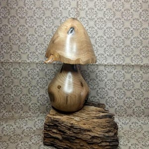 Pode incluir: Uma escultura de cogumelo em madeira torneada à mão com um grão castanho-claro liso e polido, apresentando um pequeno nó natural no chapéu e um caule arredondado, assente sobre uma peça retangular de casca de madeira escura e rugosa.