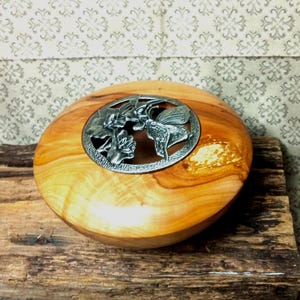 Peut inclure: Un bol rond en bois poli présentant une incrustation circulaire en métal argenté au centre. L'insert métallique affiche un motif en relief détaillé représentant un papillon et des éléments floraux, contrastant avec le grain du bois doré.