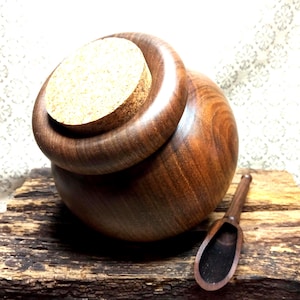 Può includere: Un barattolo sferico in legno marrone scuro con una finitura liscia e lucida e un bordo spesso e arrotondato. Il barattolo è sigillato con un tappo di sughero circolare color marrone chiaro. Accanto al barattolo c'è un piccolo cucchiaio in legno.