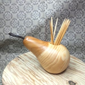Pode incluir: Um porta-palitos de madeira em forma de pêra com um caule castanho escuro. O suporte é feito de madeira de cor clara e contém vários palitos de madeira. O suporte está sobre uma superfície redonda de madeira contra um fundo padronizado.