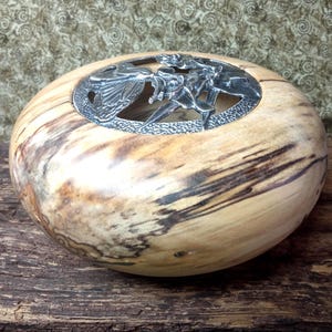 Peut inclure: Un bol en bois poli et arrondi présentant un grain naturel beige clair avec des stries brun foncé. L'ouverture supérieure est recouverte d'un insert circulaire en métal argenté orné d'un motif découpé complexe de papillon et de fleurs.