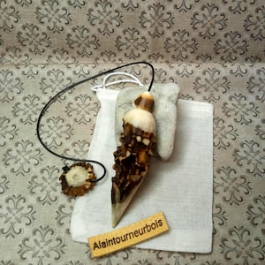 May include: A pointed, cone-shaped pendant crafted from dark brown and cream-colored wood with a textured, natural bark-like surface. It hangs from a thin black cord attached to a small, circular wooden bead. A small rectangular tag reads Alaintourneurbois.