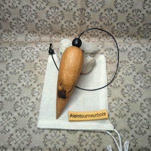 Op de afbeelding: Een druppelvormige houten pendel met een natuurlijke nerf en een donkere knoest, bevestigd aan een zwart koord met een kleine zwarte kraal. Een rechthoekig label met de tekst "Alaintourneurbois" ligt op een klein wit stoffen zakje.