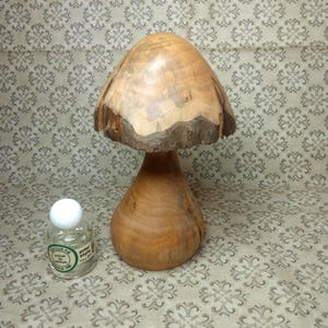 Peut inclure: Une sculpture de champignon en bois sculpté à la main avec un chapeau lisse brun clair et un bord en écorce naturelle, placée à côté d'un petit flacon de parfum en verre transparent avec un bouchon blanc et l'étiquette Senteur Pin Extrait de Parfum Prim Style.