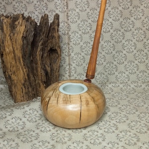 Op de afbeelding: Een houten kaarsenhouder met een lichtbruine afwerking en een witte keramische inzet. De houder is rond en heeft een glad oppervlak. Een houten pen is aan de bovenkant van de houder bevestigd.