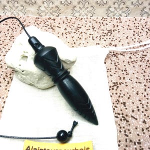 May include: A black wooden pendulum with a pointed tip and carved geometric details, attached to a black cord with a single black bead. A small yellow rectangular tag with the text "Alaintourneurbois" is attached to the cord.