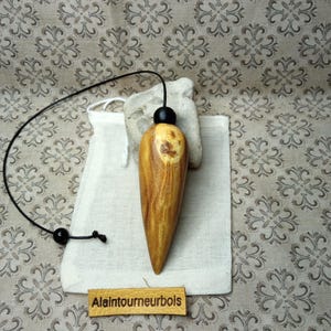 Può includere: Un pendolo in legno levigato a forma di goccia con venature visibili, fissato a un cordino nero con una perlina nera. Poggia su un piccolo sacchetto di tessuto bianco, accompagnato da un'etichetta in pelle color cuoio con la scritta Alaintourneurbols.