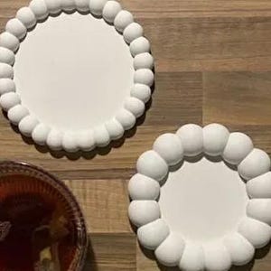 Pode incluir: Duas bases redondas brancas com borda de contas são exibidas em uma superfície de madeira. As bases têm um design simples e elegante. Um copo de chá e uma flor branca também são visíveis.