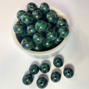 May include: A collection of green and black plaid wooden beads, some inside a white bowl and others scattered on a white surface. The beads are round with a hole drilled through the center, suitable for stringing.
