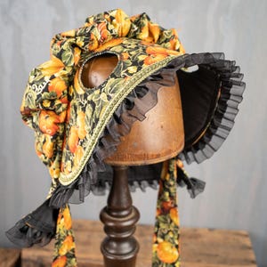 May include: A black and yellow fabric bonnet with a ruffled trim and a floral pattern featuring pumpkins. The bonnet is on a wooden display stand.