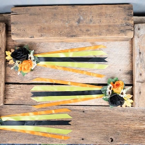 May include: Three black and orange flower arrangements with black and yellow ribbon streamers on a rustic wooden background.
