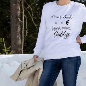 May include: A white sweatshirt with the text "Never doubt your inner Goddess" in black script. The design includes an arrow and a crescent moon. The person is wearing blue jeans and holding a tan jacket.