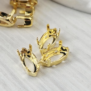 May include: Several gold-colored jewelry settings are displayed on a white surface. The settings have an oval shape with four prongs and a base with a rectangular opening. The metal has a shiny finish.