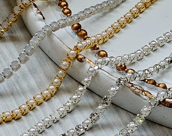 Puede incluir: Primer plano de varias hebras de cadena decorativa. Las cadenas varían en color, incluyendo oro y plata, y presentan pequeños adornos redondos que se asemejan a perlas y cristales. Las cadenas están dispuestas diagonalmente sobre una superficie blanca.