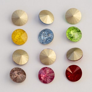 May include: Assortment of twelve round, flat-backed rhinestones in various colors, including gold, yellow, blue, green, red, and brown. The rhinestones have a faceted design and are arranged in a grid pattern.