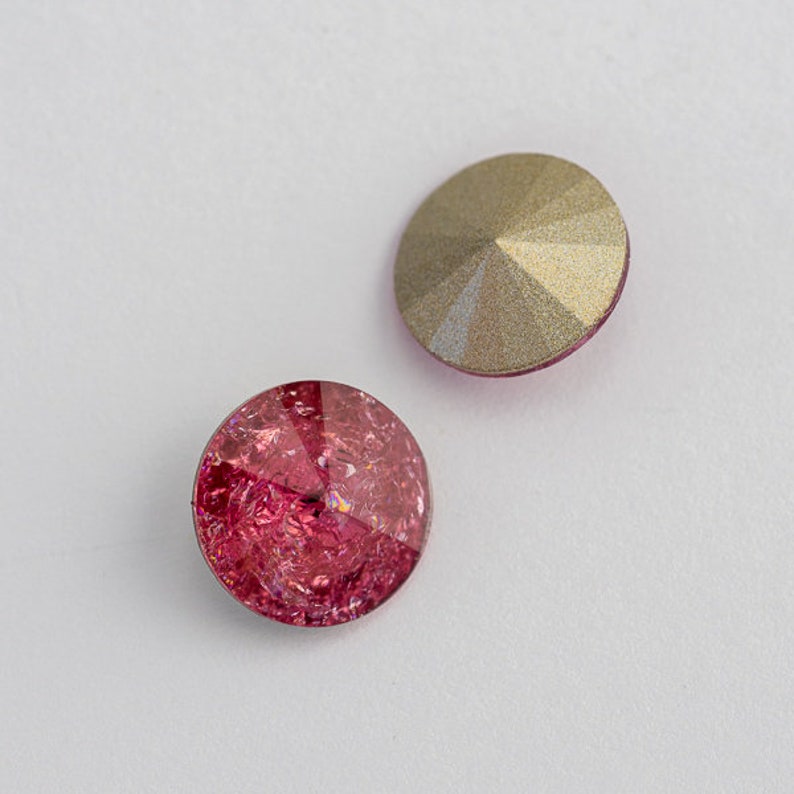 May include: Two round, flat-backed rhinestones. One is a clear gold color and the other is a deep pink with a glittery finish.