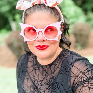 Puede incluir: Una persona lleva gafas de sol rosas con forma de murci&eacute;lago con lentes rojas y una diadema a juego con un estampado de murci&eacute;lago. Lleva una camiseta negra con un dise&ntilde;o de telara&ntilde;a. Sus labios est&aacute;n pintados de rojo.