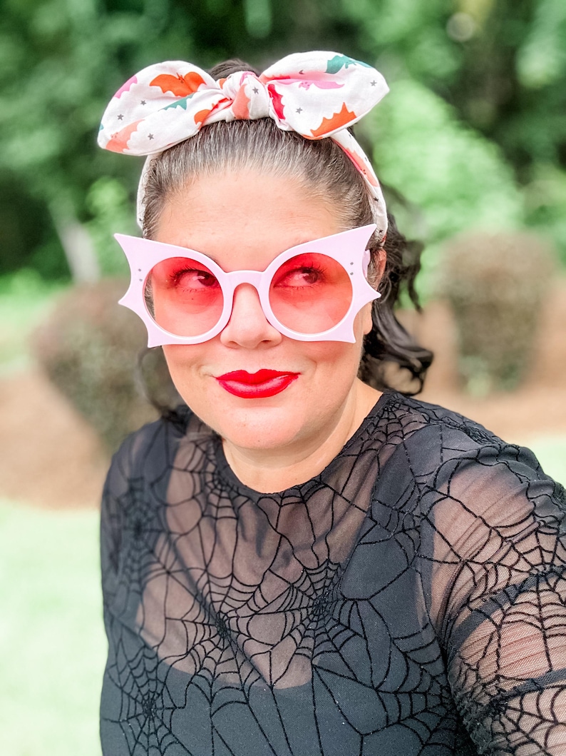 Puede incluir: Una persona lleva gafas de sol rosas con forma de murci&eacute;lago con lentes rojas y una diadema a juego con un estampado de murci&eacute;lago. Lleva una camiseta negra con un dise&ntilde;o de telara&ntilde;a. Sus labios est&aacute;n pintados de rojo.