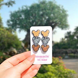 May include: A pair of dangle earrings featuring a three-tiered design. The top tier has a gold glitter finish, the middle tier has a zebra print, and the bottom tier has a tiger stripe pattern. The earrings are held on a white card with the text "Mommin' with the Myuse".