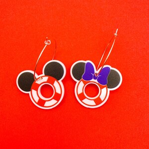Puede incluir: Un par de pendientes de aro con un salvavidas rojo y blanco y orejas de Mickey Mouse negras. Un pendiente tiene un lazo morado en las orejas.