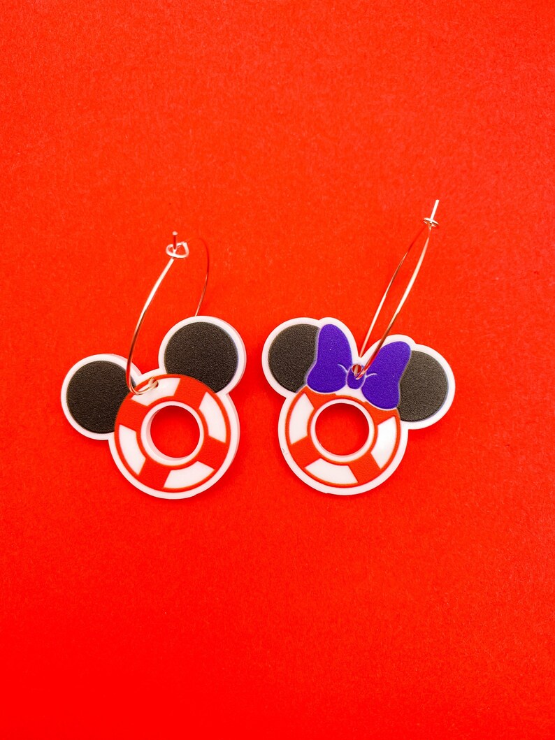 Puede incluir: Un par de pendientes de aro con un salvavidas rojo y blanco y orejas de Mickey Mouse negras. Un pendiente tiene un lazo morado en las orejas.