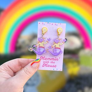 May include: Acrylic earrings featuring a purple dragon character with a yellow shirt and lightbulb accents. The earrings are held in front of a rainbow and a sign that says "Mommin' with the Mouse".