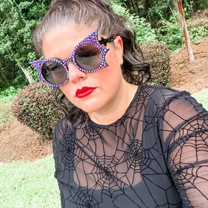 Puede incluir: Una persona con gafas de sol negras con dise&ntilde;o de ala de murci&eacute;lago y una camiseta de malla negra con un patr&oacute;n de telara&ntilde;a. Las gafas de sol tienen un ribete de joyas moradas y azules. La persona tiene pintalabios rojo.