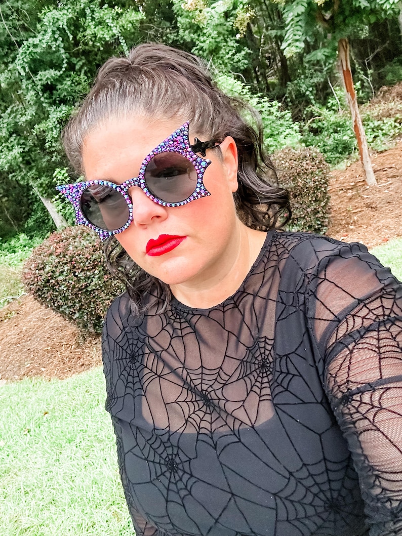 Puede incluir: Una persona con gafas de sol negras con dise&ntilde;o de ala de murci&eacute;lago y una camiseta de malla negra con un patr&oacute;n de telara&ntilde;a. Las gafas de sol tienen un ribete de joyas moradas y azules. La persona tiene pintalabios rojo.