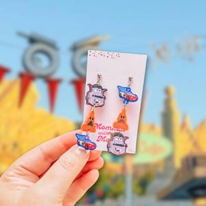Puede incluir: Pendientes colgantes con personajes de dibujos animados de coches y conos de tráfico naranjas. Los pendientes están sujetos a una tarjeta blanca con el texto "Moments with Ma". El fondo es una escena exterior borrosa.