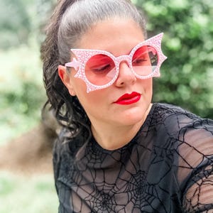 Puede incluir: Gafas de sol rosas con dise&ntilde;o de ala de murci&eacute;lago y lentes rosas. Las monturas est&aacute;n incrustadas con peque&ntilde;os pedrer&iacute;a brillantes. La persona lleva una camiseta negra con un estampado de telara&ntilde;a.