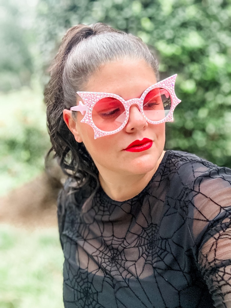 Puede incluir: Gafas de sol rosas con dise&ntilde;o de ala de murci&eacute;lago y lentes rosas. Las monturas est&aacute;n incrustadas con peque&ntilde;os pedrer&iacute;a brillantes. La persona lleva una camiseta negra con un estampado de telara&ntilde;a.