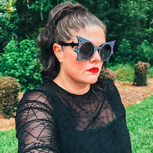 Puede incluir: Gafas de sol negras con dise&ntilde;o de ala de murci&eacute;lago y un patr&oacute;n de puntos de colores. Las gafas de sol se llevan con una camiseta negra de manga larga con un dise&ntilde;o de telara&ntilde;a. La persona lleva pintalabios rojo.