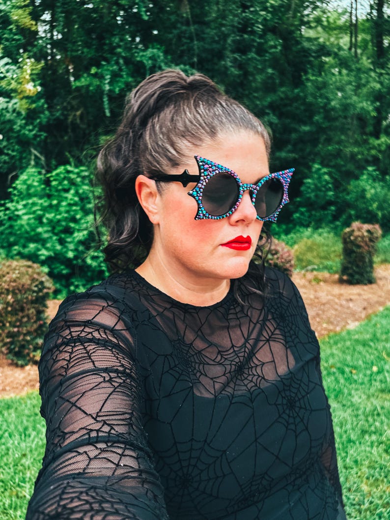 Puede incluir: Gafas de sol negras con dise&ntilde;o de ala de murci&eacute;lago y un patr&oacute;n de puntos de colores. Las gafas de sol se llevan con una camiseta negra de manga larga con un dise&ntilde;o de telara&ntilde;a. La persona lleva pintalabios rojo.