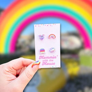 May include: A set of four colorful stud earrings on a white card, held by a hand with rainbow nail art. The earrings feature a purple creature, a rainbow, a colorful egg, and a flower. The card reads "Mommin' with the Mouse".