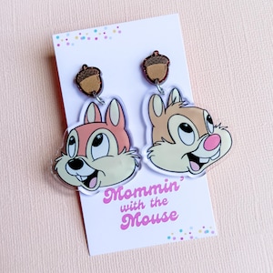 Puede incluir: Pendientes colgantes con caras de ardillas de dibujos animados, una rosa y otra marrón claro, con adornos de bellotas. Los pendientes se muestran en una tarjeta blanca con el texto "Mommin' with the Mouse" en rosa.