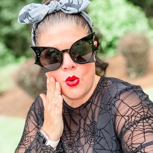 Puede incluir: Gafas de sol negras con forma de murci&eacute;lago y lentes redondas. Las gafas se usan con una diadema gris con un patr&oacute;n de murci&eacute;lago. La persona lleva una camiseta negra con un dise&ntilde;o de telara&ntilde;a. El l&aacute;piz labial rojo completa el look.