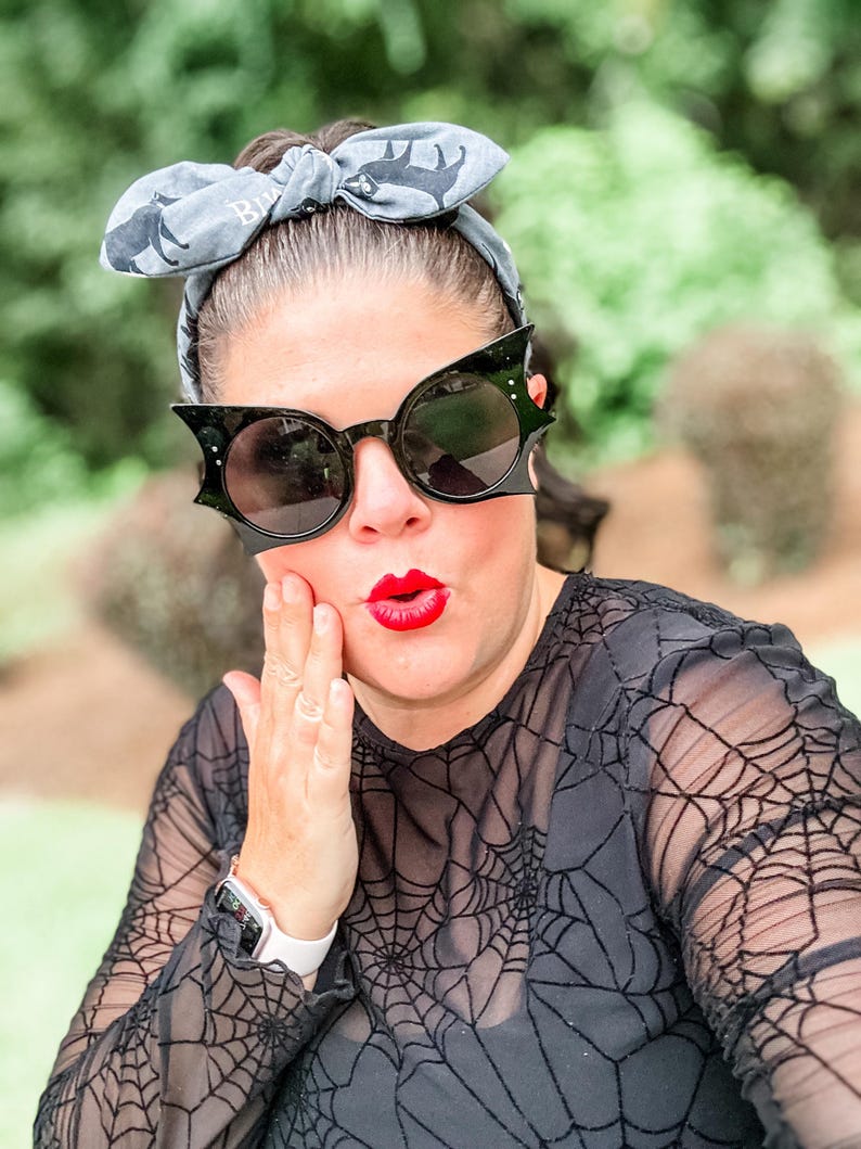 Puede incluir: Gafas de sol negras con forma de murci&eacute;lago y lentes redondas. Las gafas se usan con una diadema gris con un patr&oacute;n de murci&eacute;lago. La persona lleva una camiseta negra con un dise&ntilde;o de telara&ntilde;a. El l&aacute;piz labial rojo completa el look.