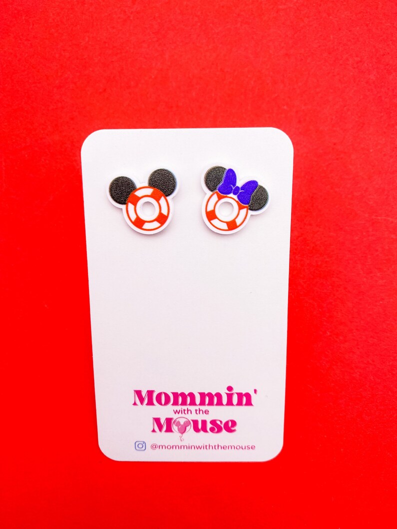 Puede incluir: Un par de pendientes de bot&oacute;n blancos con orejas de Mickey Mouse rojas, blancas y negras y un salvavidas rojo, blanco y negro. Los pendientes est&aacute;n en una tarjeta blanca con el texto "Mommin' with the Mouse" y el nombre de usuario de Instagram "@momminwiththemouse".