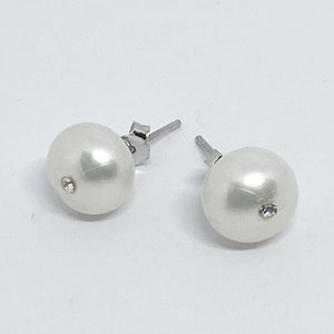 May include: A pair of white pearl stud earrings with a small, clear crystal accent on each. The earrings have silver-colored posts and are set against a white background. The pearls are round and have a smooth, glossy finish.