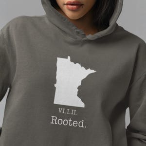 Könnte beinhalten: Grauer Kapuzenpullover mit weißer Umrisszeichnung des Bundesstaates Minnesota. Unter der Umrisszeichnung stehen die römischen Ziffern VI.I.II. und das Wort „Rooted“. Der Hoodie hat eine Kapuze und eine lockere Passform.