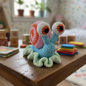 Puede incluir: Un juguete de caracol hecho a mano con ganchillo con una concha a rayas rosa y naranja, cuerpo azul y ojos blancos grandes con pupilas negras. El caracol está sentado en una mesa de madera. El fondo incluye una ventana, plantas y juguetes para niños.